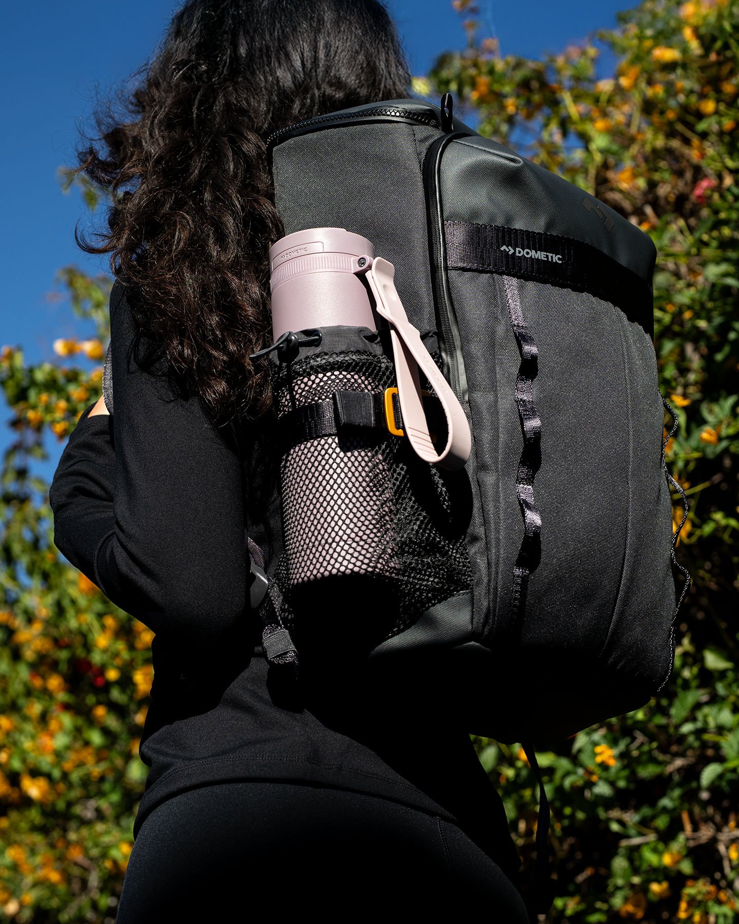 Person wearing a black Dometic rucksack outdoors with a pink insulated bottle in the side mesh pocket.
