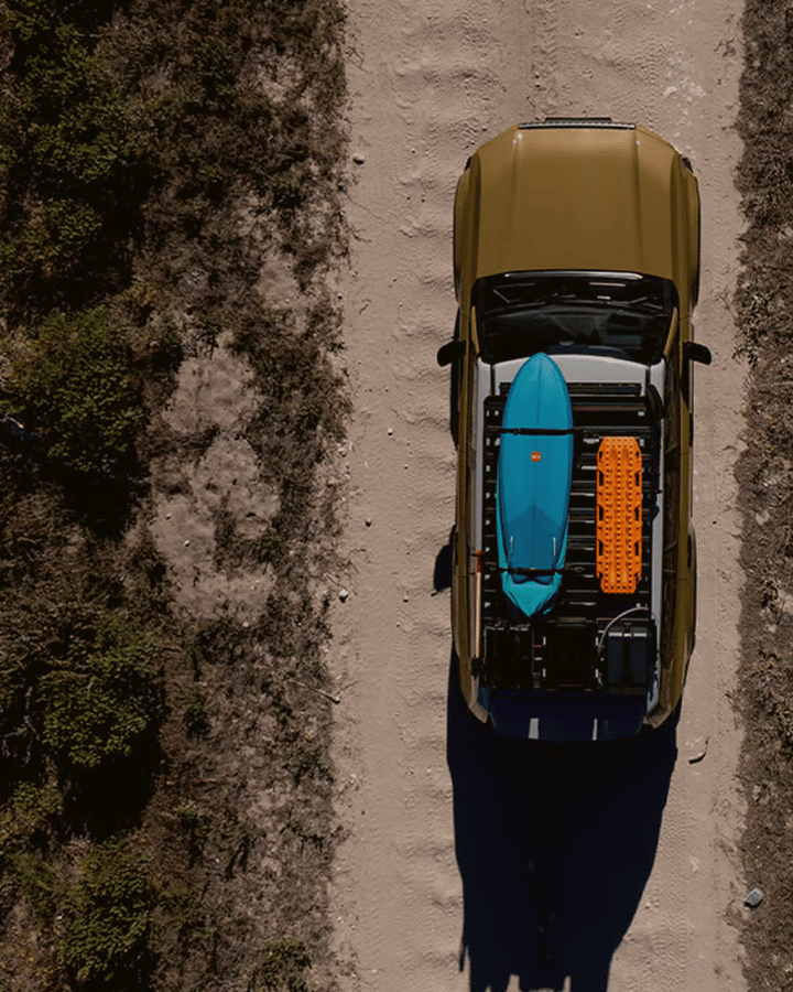 Aerial top‑down view of a vehicle on a dirt road with a roof rack carrying a surfboard and recovery board.