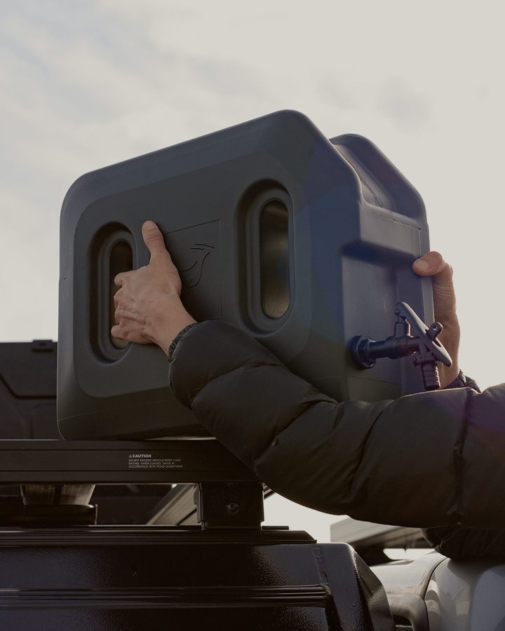 A person lifting a portable water or fuel container onto a roof rack platform.