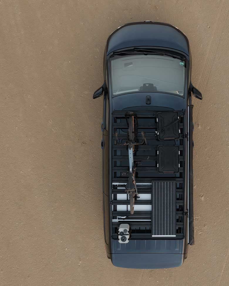 Aerial top‑down view of a vehicle with mounted roof‑rack gear and a secured bicycle on a sandy surface.