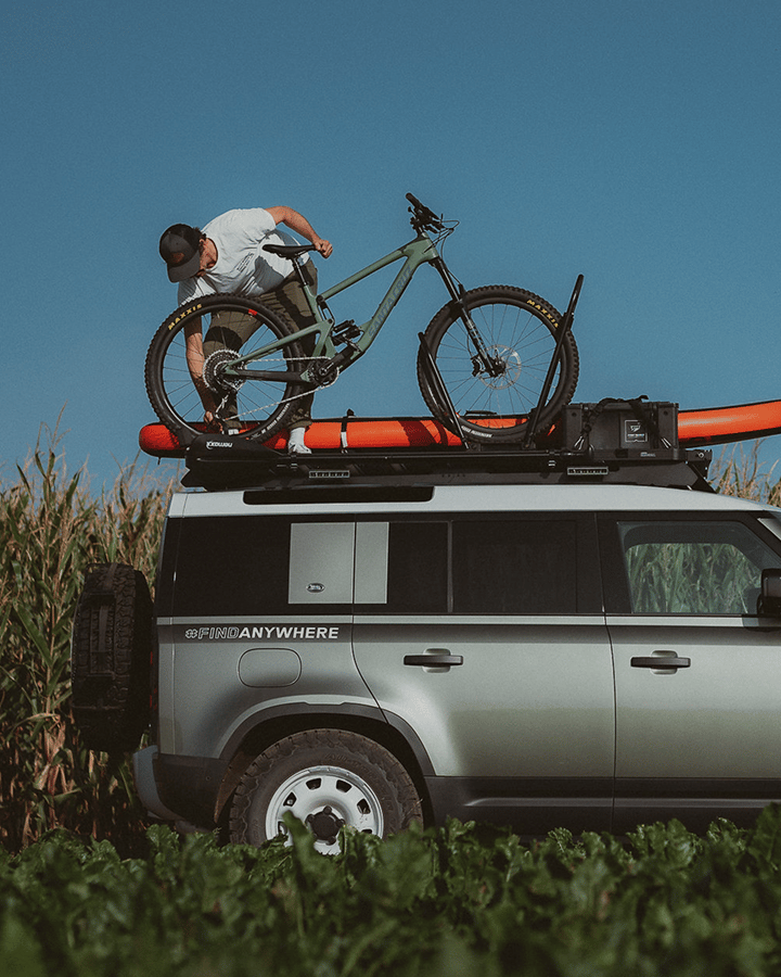 A person loading a bicycle onto a roof rack carrying a kayak and gear on a vehicle beside tall vegetation.