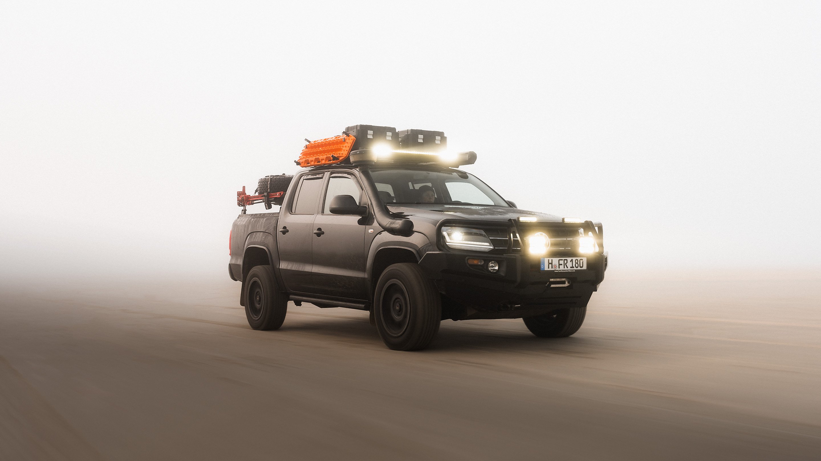 Pickup truck equipped with a roof rack, LED light bar, recovery boards and storage boxes driving in dusty off-road conditions