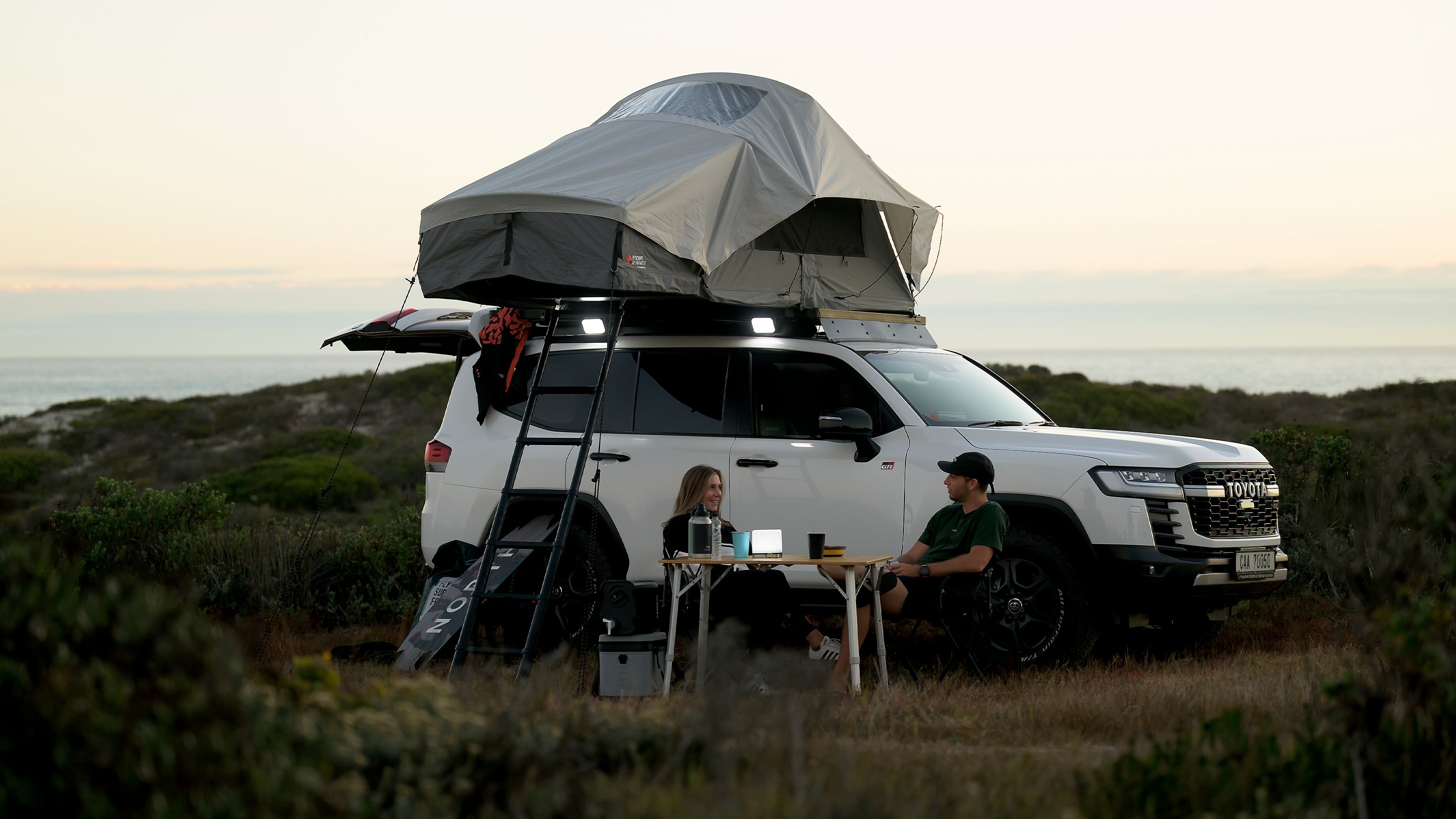 White vehicle parked near the coastline at sunset with a Dometic Front Runner Series Feather‑Lite II roof tent set up, ladder in place, and a camping table next to the vehicle.