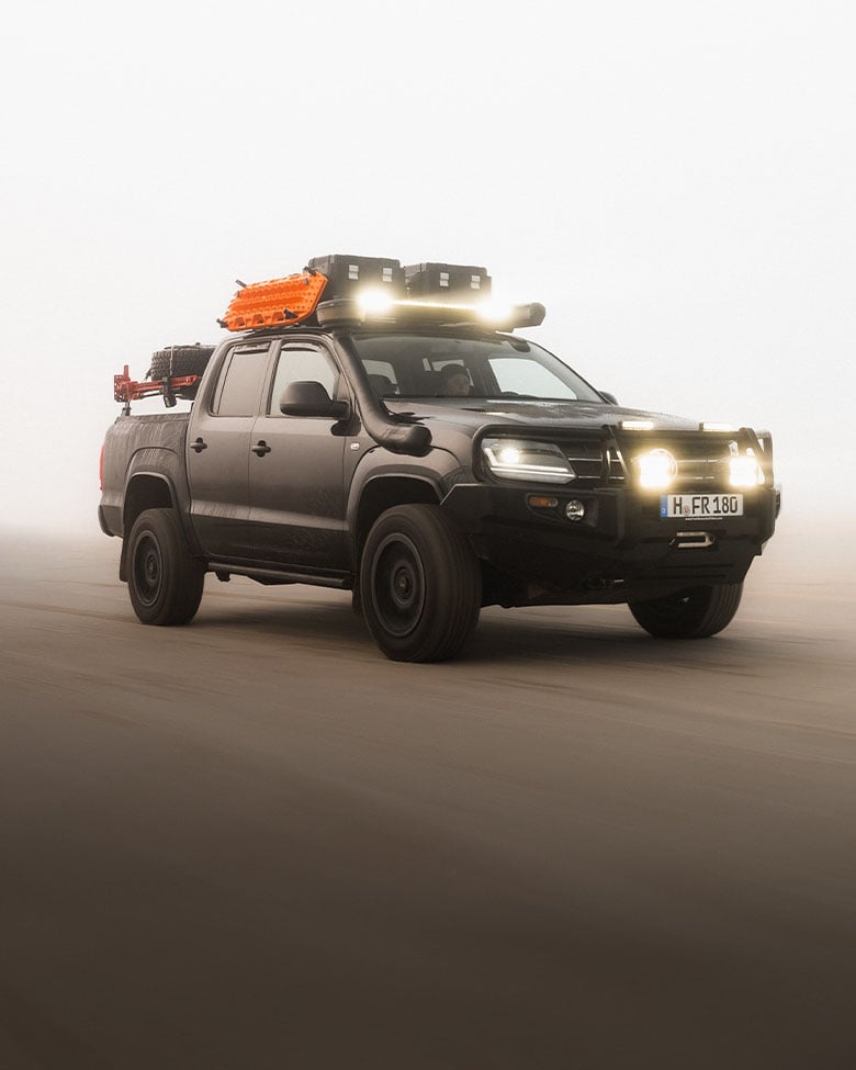 Pickup truck equipped with a roof rack, LED light bar, recovery boards and storage boxes driving in dusty off-road conditions