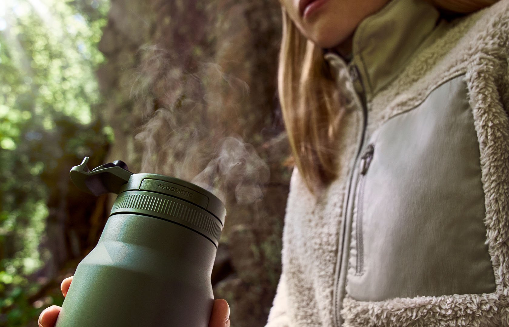 Green insulated thermo bottle with steam rising from the lid, held outdoors near a tree.