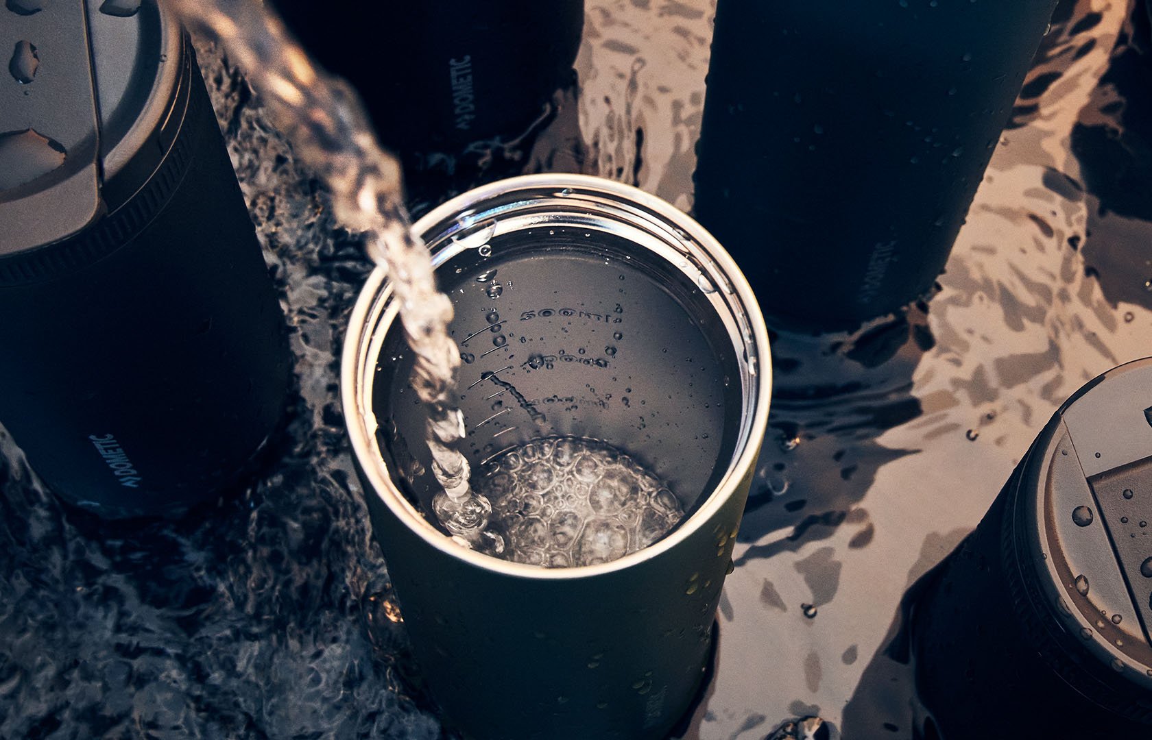 Insulated metal tumbler being filled with water in shallow outdoor water, surrounded by other drinkware.