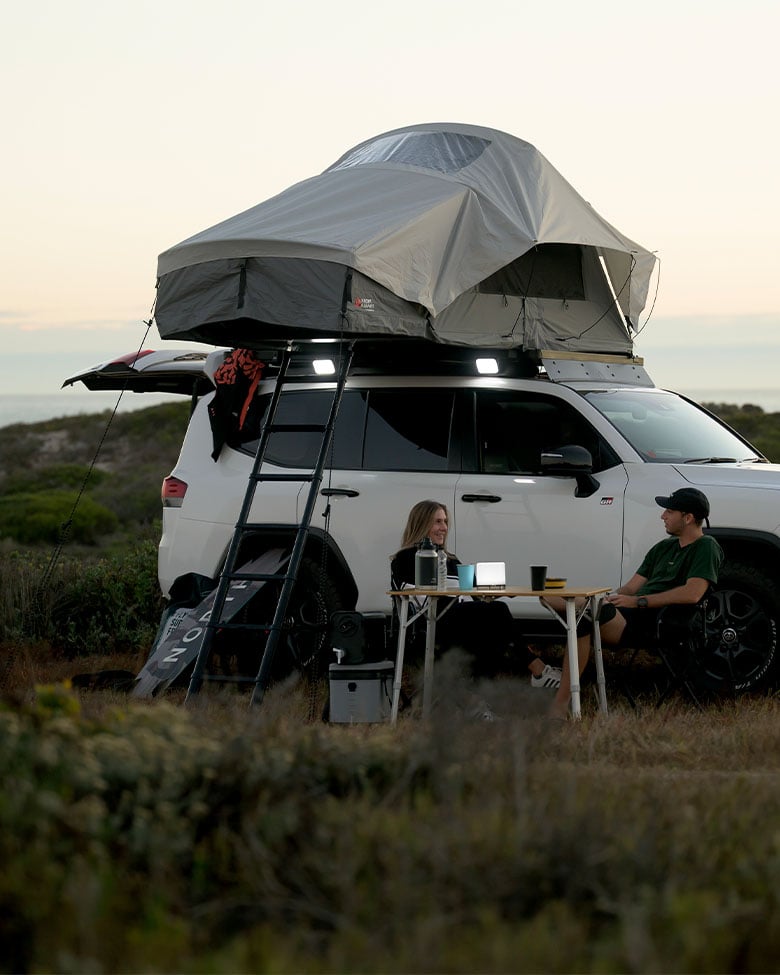 White vehicle parked near the coastline at sunset with a Dometic Front Runner Series Feather‑Lite II roof tent set up, ladder in place, and a camping table next to the vehicle.