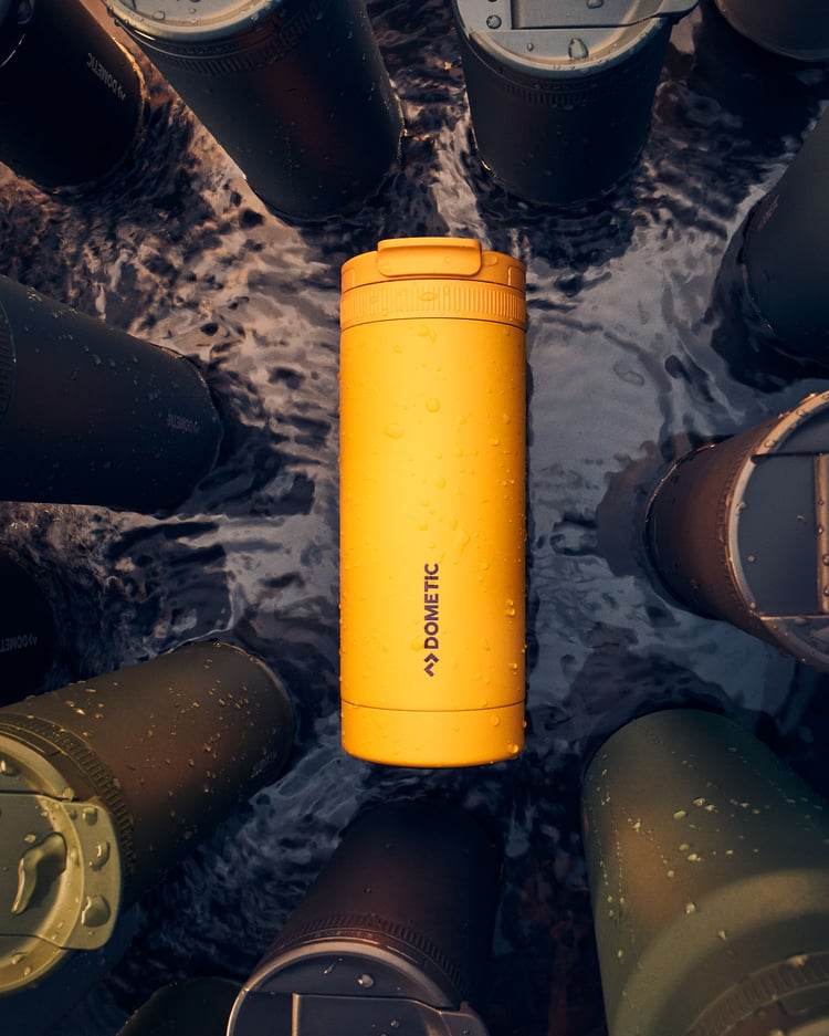 Yellow insulated thermo tumbler with water droplets surrounded by outdoor drinkware in shallow water.